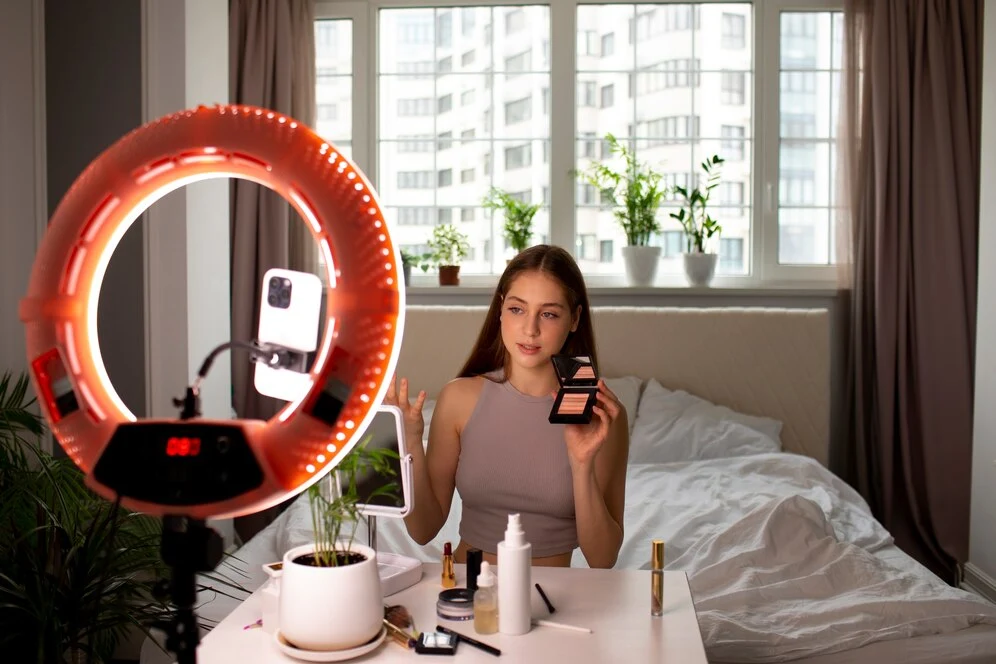 Young woman sitting at a table with makeup products, recording a beauty tutorial with a smartphone mounted on a ring light, in a casual bedroom setting with a window and potted plants