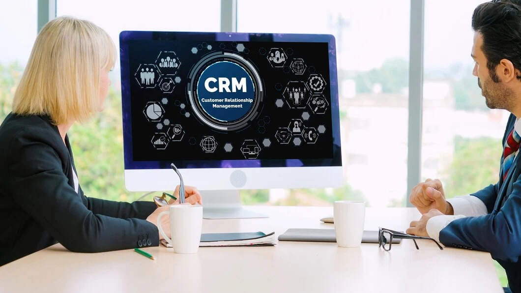 A corporate setting with two colleagues discussing a Customer Relationship Management system as seen on the computer screen,